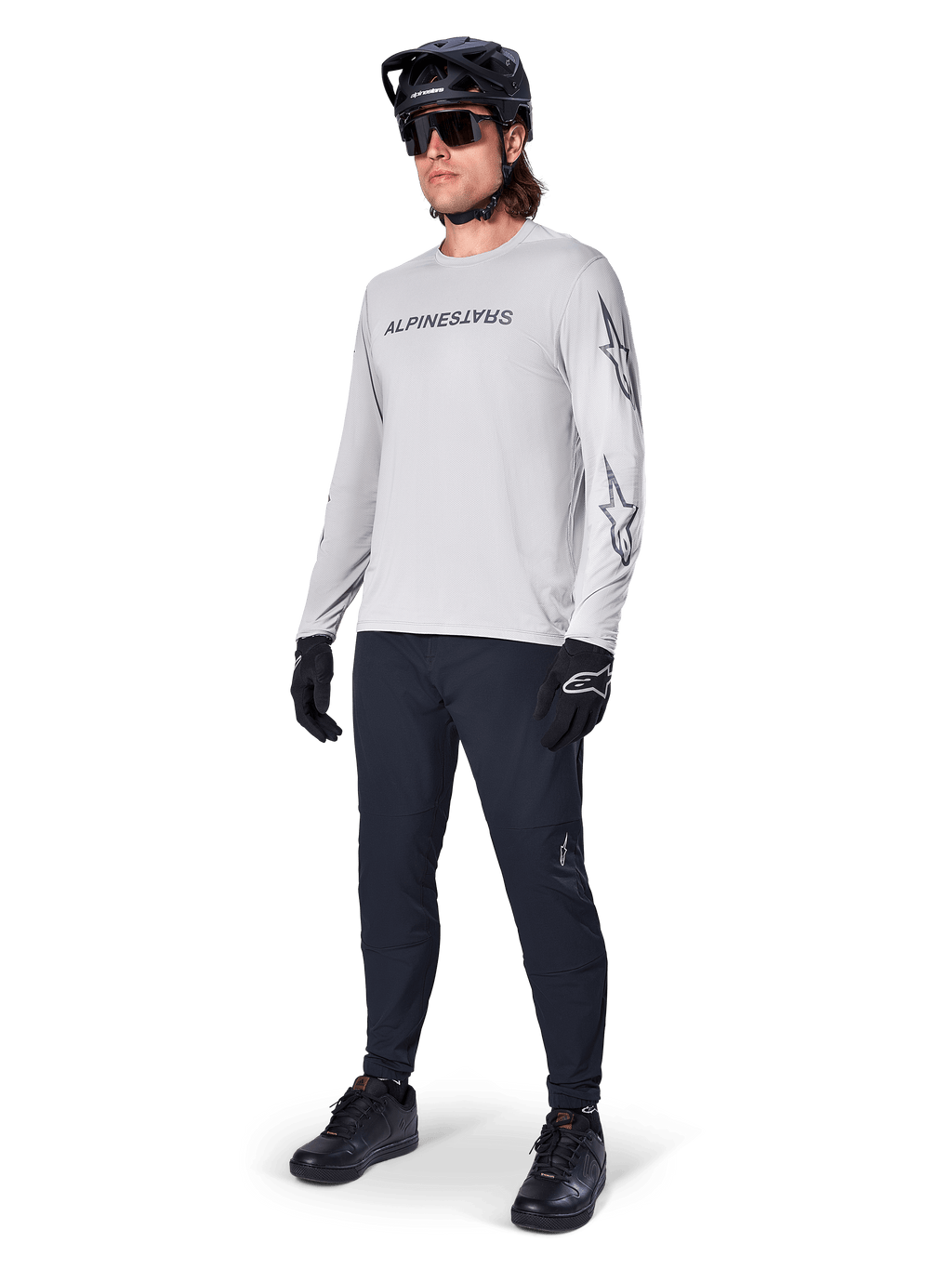 Alpinestars A-Dura Switch Long Sleeve Jersey, jasnoszara, noszona przez rowerzystę z czarnymi spodniami MTB i kaskiem, czarny logotyp na piersi i grafiki gwiazd na rękawach, profesjonalna odzież do jazdy terenowej