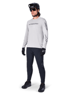 Alpinestars A-Dura Switch Long Sleeve Jersey, jasnoszara, noszona przez rowerzystę z czarnymi spodniami MTB i kaskiem, czarny logotyp na piersi i grafiki gwiazd na rękawach, profesjonalna odzież do jazdy terenowej