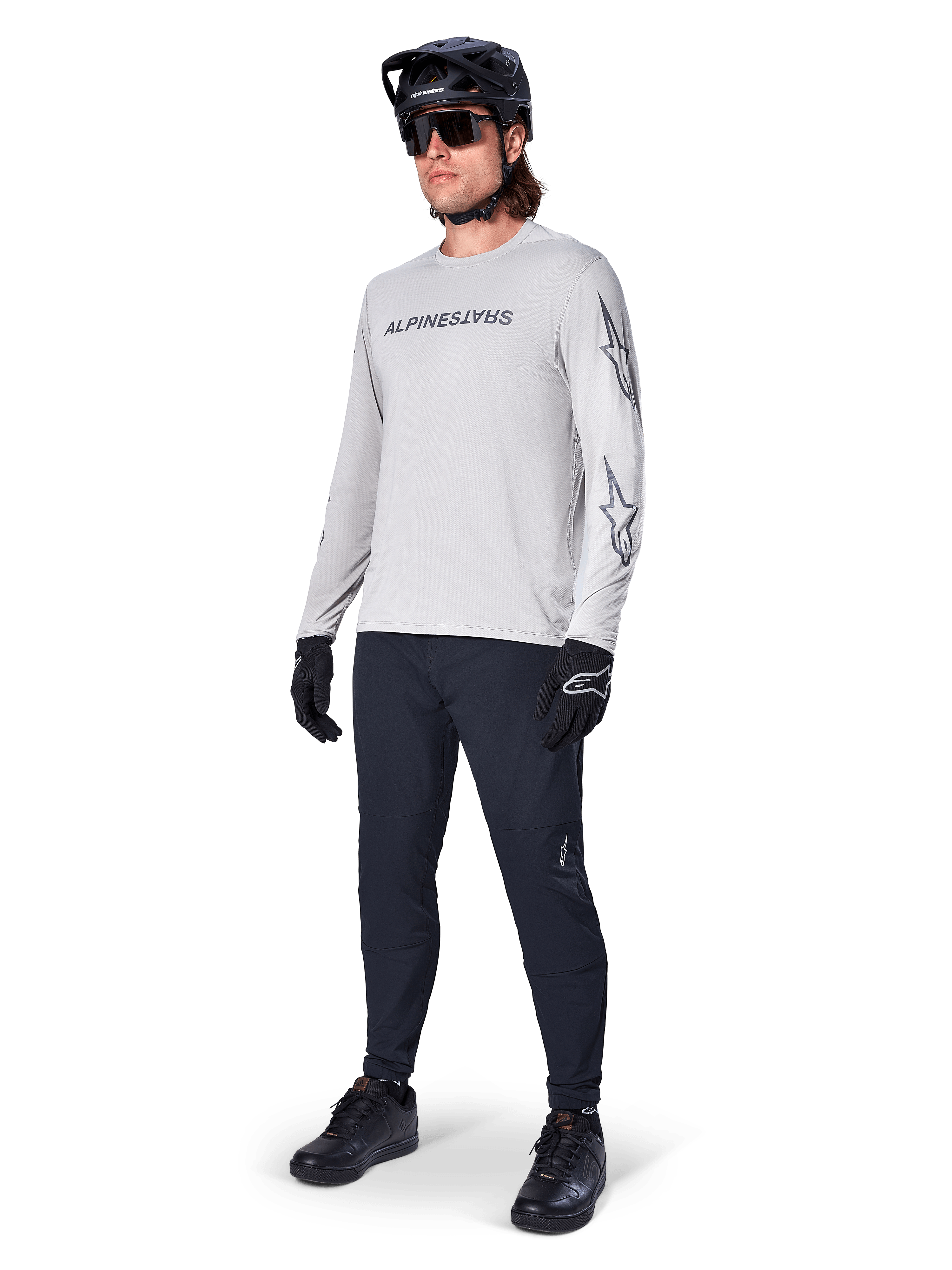 Alpinestars A-Dura Switch Long Sleeve Jersey, jasnoszara, noszona przez rowerzystę z czarnymi spodniami MTB i kaskiem, czarny logotyp na piersi i grafiki gwiazd na rękawach, profesjonalna odzież do jazdy terenowej