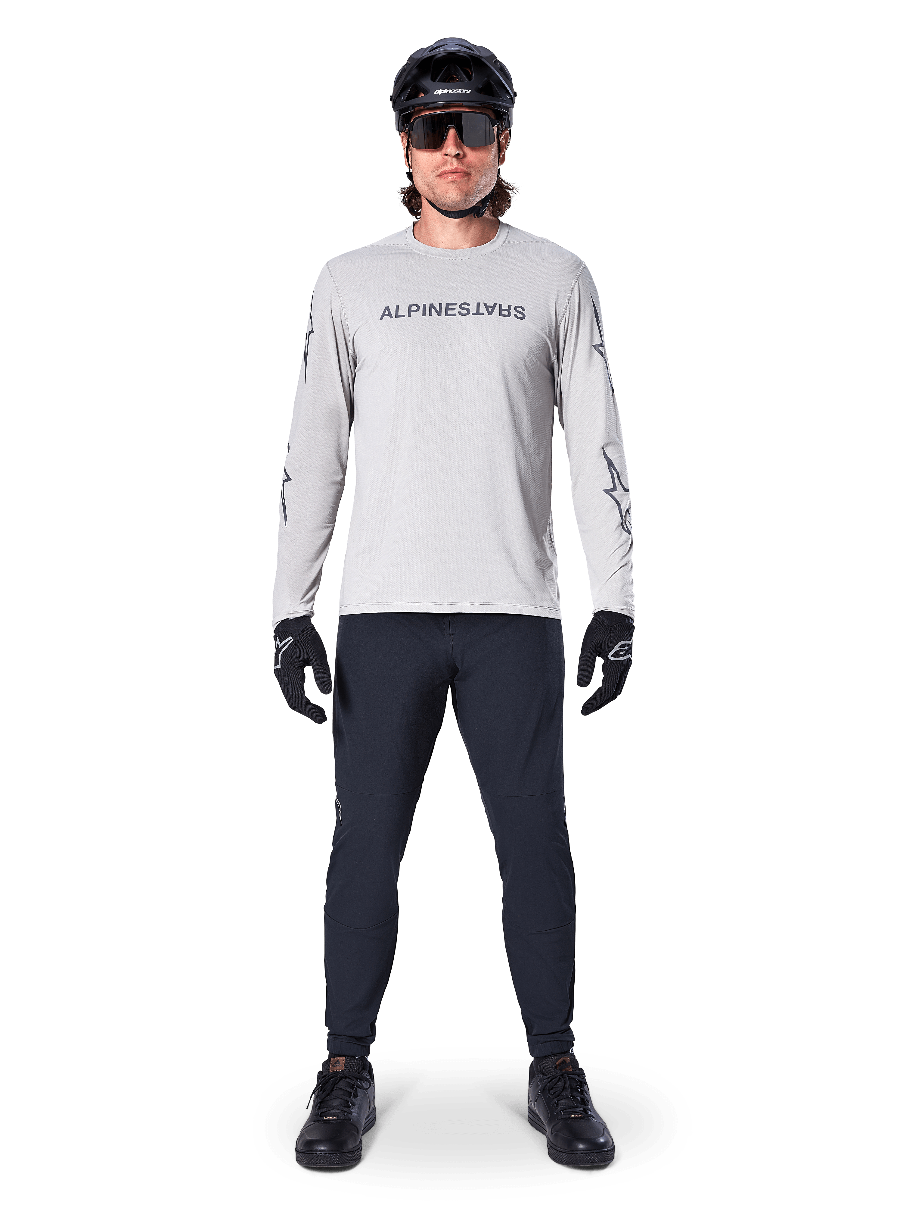 Koszulka Alpinestars A-Dura Switch z długim rękawem w kolorze jasnoszarym, ze stylizowanym logotypem na piersi, noszona z czarnymi spodniami, kaskiem i rękawiczkami, profesjonalna odzież do kolarstwa górskiego