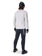 Alpinestars A-Dura Switch Long Sleeve Jersey, jasnoszara koszulka rowerowa, widok z tyłu na rowerzystę w czarnych spodniach, kasku i rękawicach, odzież MTB o wysokiej wydajności z wydłużonym tyłem