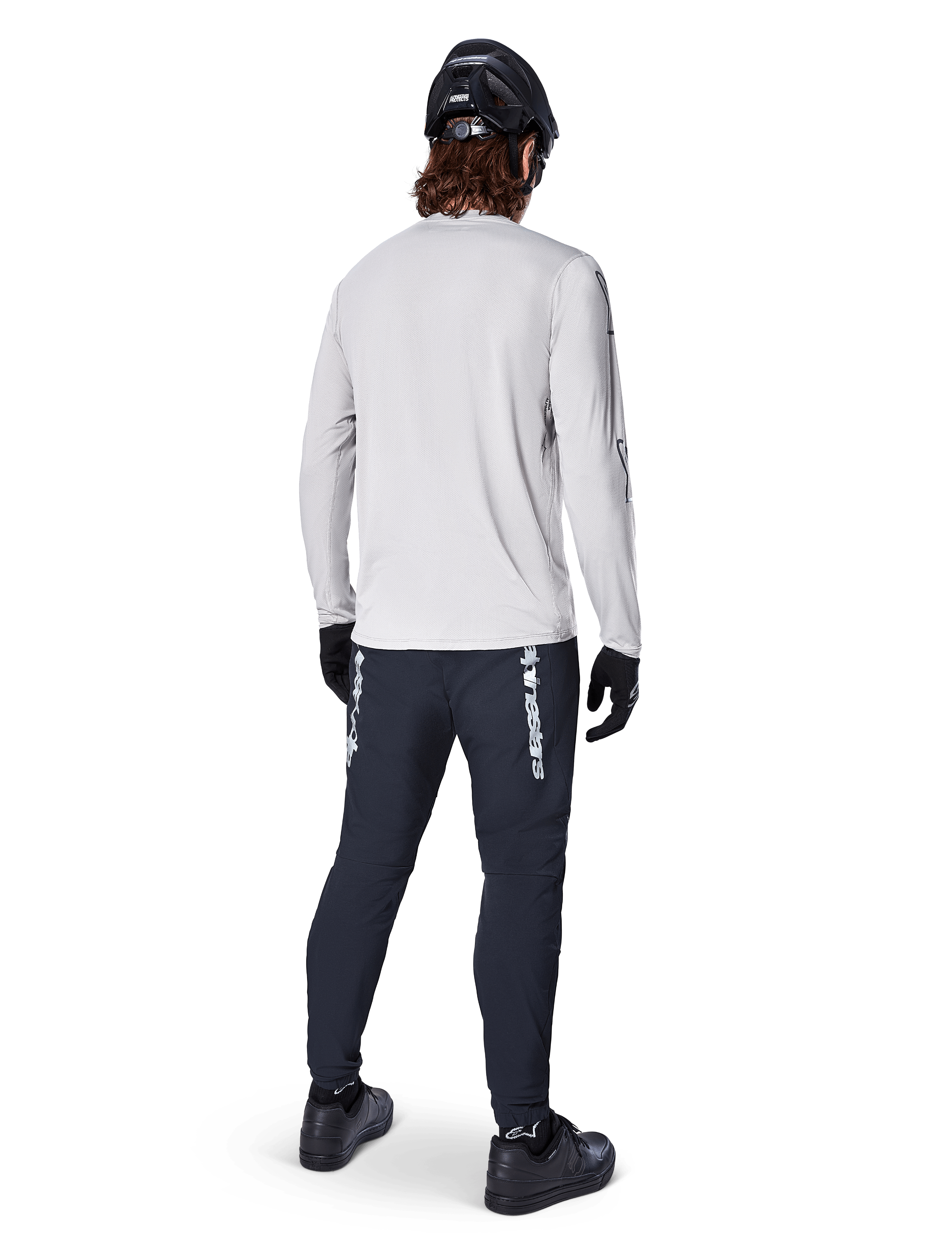 Alpinestars A-Dura Switch Long Sleeve Jersey, jasnoszara koszulka rowerowa, widok z tyłu na rowerzystę w czarnych spodniach, kasku i rękawicach, odzież MTB o wysokiej wydajności z wydłużonym tyłem