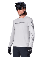 Alpinestars A-Dura Switch Long Sleeve Jersey, koszulka na rower górski, jasnoszara, czarny nadruk z nazwą marki na piersi i logo z gwiazdą na rękawach, noszona z czarnym kaskiem rowerowym MTB, ciemnymi okularami i czarnymi rękawiczkami, sportowa odzież do kolarstwa terenowego