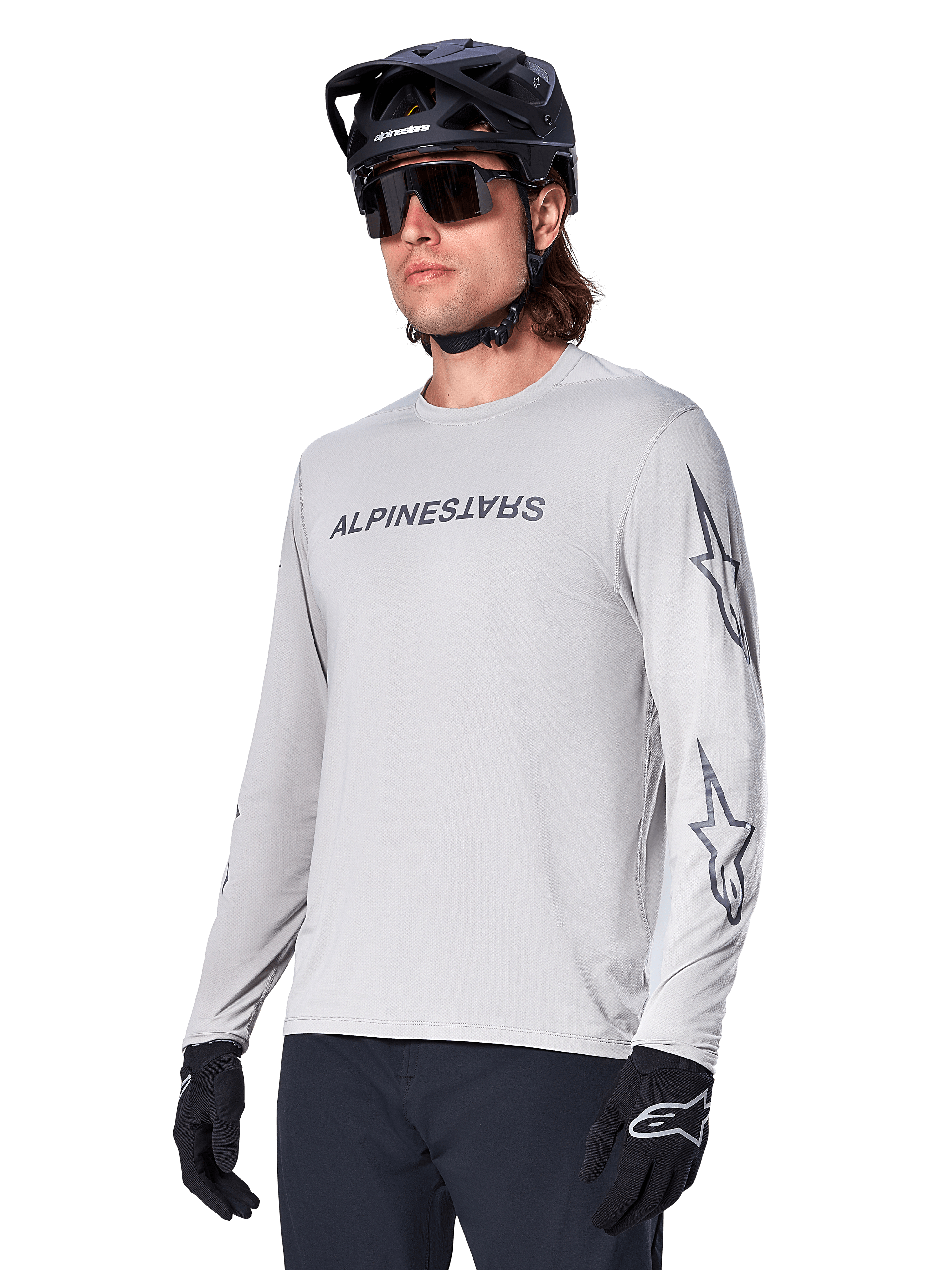 Alpinestars A-Dura Switch Long Sleeve Jersey, koszulka na rower górski, jasnoszara, czarny nadruk z nazwą marki na piersi i logo z gwiazdą na rękawach, noszona z czarnym kaskiem rowerowym MTB, ciemnymi okularami i czarnymi rękawiczkami, sportowa odzież do kolarstwa terenowego