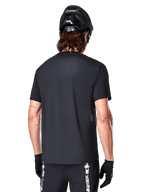 Alpinestars A-Dura Switch Short Sleeve Jersey, czarna, widok z tyłu na modelu w czarnym kasku rowerowym i spodenkach MTB, podkreślający regularny krój i przedłużony tył do jazdy na rowerze górskim