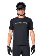 Rowerzysta górski w koszulce Alpinestars A-Dura Switch z krótkim rękawem w kolorze czarnym, z białym logo na piersi, stylizacja z czarnym kaskiem, dużymi okularami przeciwsłonecznymi i czarnymi rękawiczkami, profesjonalna odzież rowerowa do jazdy po szlakach