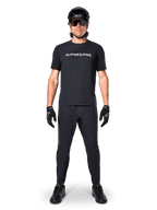 Rowerzysta górski ubrany w koszulkę Alpinestars A-Dura Switch Short Sleeve w kolorze czarnym, z białym stylizowanym logotypem na piersi, zestawioną z dopasowanymi czarnymi spodniami MTB, kaskiem i rękawiczkami, kompletny strój rowerowy gotowy na szlak