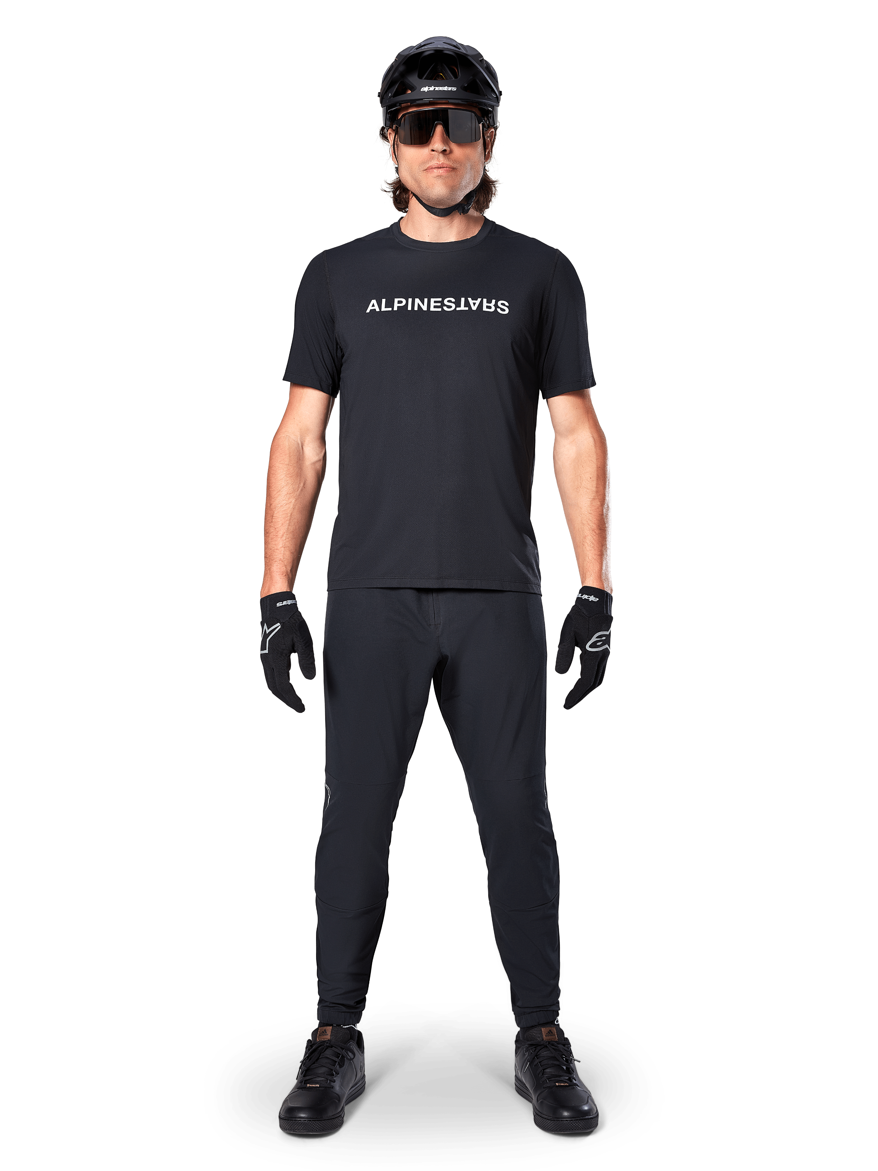 Rowerzysta górski ubrany w koszulkę Alpinestars A-Dura Switch Short Sleeve w kolorze czarnym, z białym stylizowanym logotypem na piersi, zestawioną z dopasowanymi czarnymi spodniami MTB, kaskiem i rękawiczkami, kompletny strój rowerowy gotowy na szlak