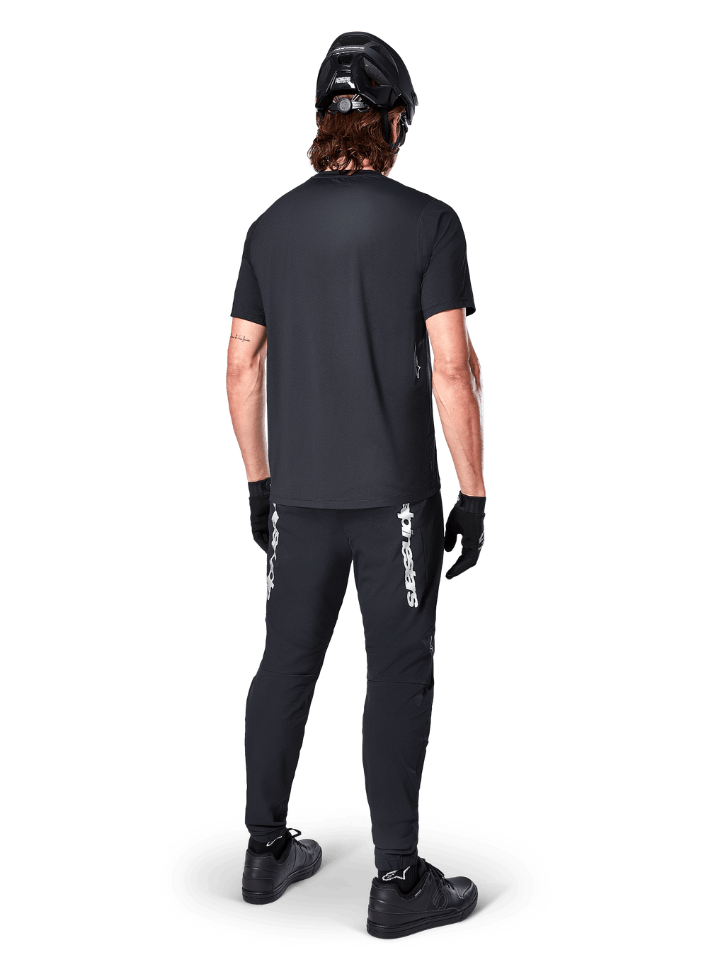 Alpinestars A-Dura Switch Jersey - Short Sleeve, czarna koszulka z krótkim rękawem, widok od tyłu na kolarza w kompletnym stroju MTB, czarne spodnie z białym logotypem, czarny kask i rękawice, techniczna odzież rowerowa do jazdy po szlakach