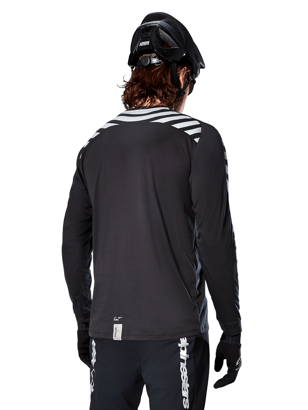 Koszulka Alpinestars A-Dura Dri Oscar z długim rękawem, Black Reflective, widok z tyłu pokazujący odblaskowe ukośne pasy na ramionach i przedłużony tył, noszona z czarnym kaskiem MTB i rękawiczkami, techniczna odzież do kolarstwa górskiego