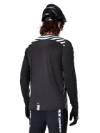 Koszulka Alpinestars A-Dura Dri Oscar z długim rękawem, Black Reflective, widok z tyłu pokazujący odblaskowe ukośne pasy na ramionach i przedłużony tył, noszona z czarnym kaskiem MTB i rękawiczkami, techniczna odzież do kolarstwa górskiego