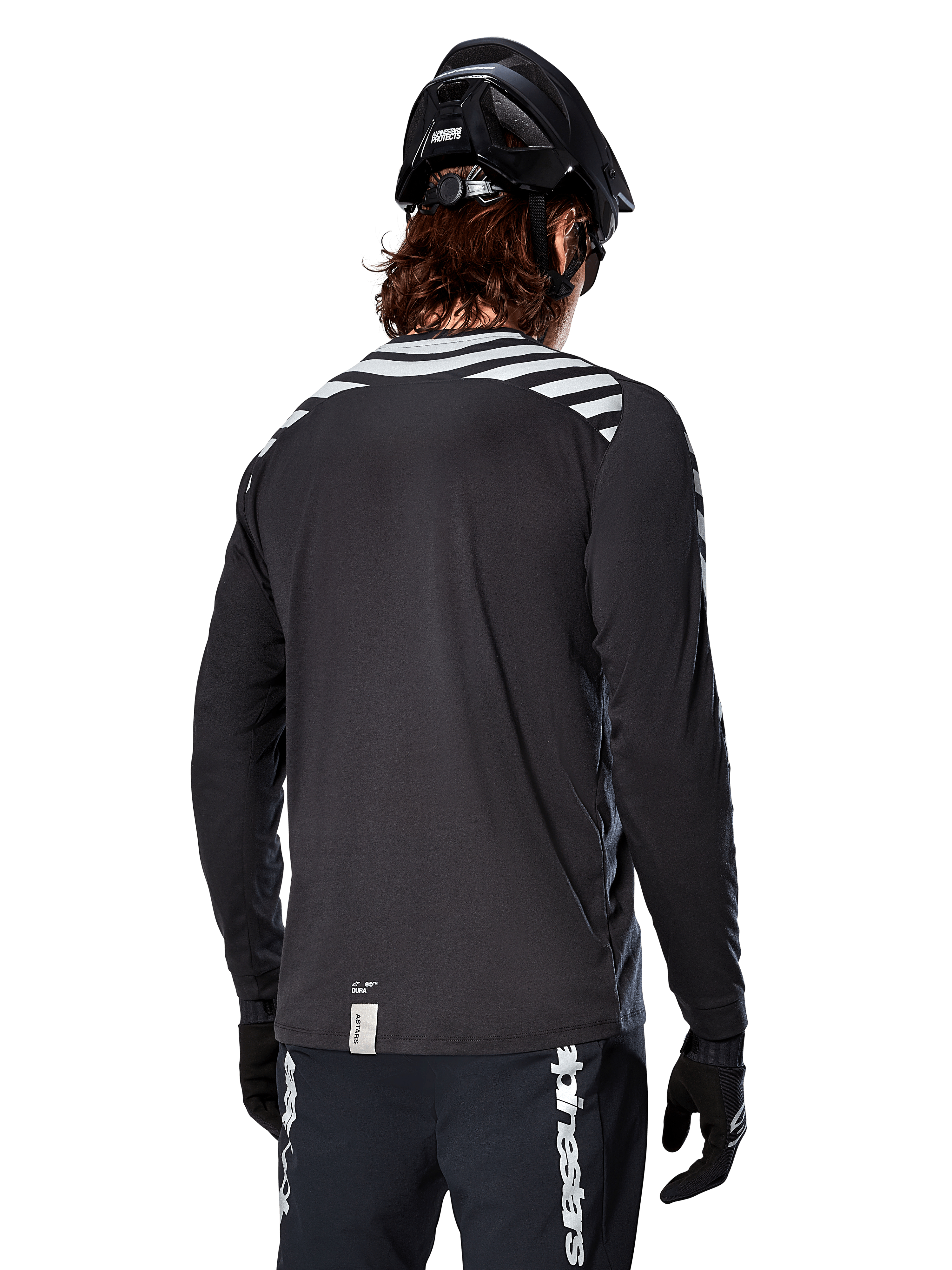 Koszulka Alpinestars A-Dura Dri Oscar z długim rękawem, Black Reflective, widok z tyłu pokazujący odblaskowe ukośne pasy na ramionach i przedłużony tył, noszona z czarnym kaskiem MTB i rękawiczkami, techniczna odzież do kolarstwa górskiego