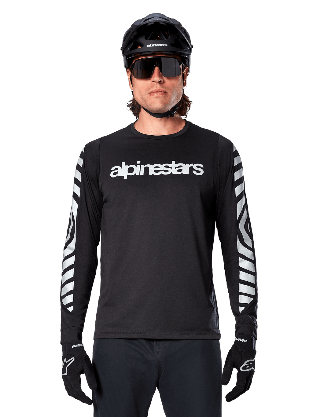 Mężczyzna w koszulce Alpinestars A-Dura Dri Oscar z długim rękawem, kolor Black Reflective, z białym napisem Alpinestars na klatce piersiowej i srebrnymi odblaskowymi grafikami na rękawach, czarny kask MTB, okulary przeciwsłoneczne i czarne rękawiczki, profesjonalna odzież rowerowa