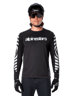 Mężczyzna w koszulce Alpinestars A-Dura Dri Oscar z długim rękawem, kolor Black Reflective, z białym napisem Alpinestars na klatce piersiowej i srebrnymi odblaskowymi grafikami na rękawach, czarny kask MTB, okulary przeciwsłoneczne i czarne rękawiczki, profesjonalna odzież rowerowa
