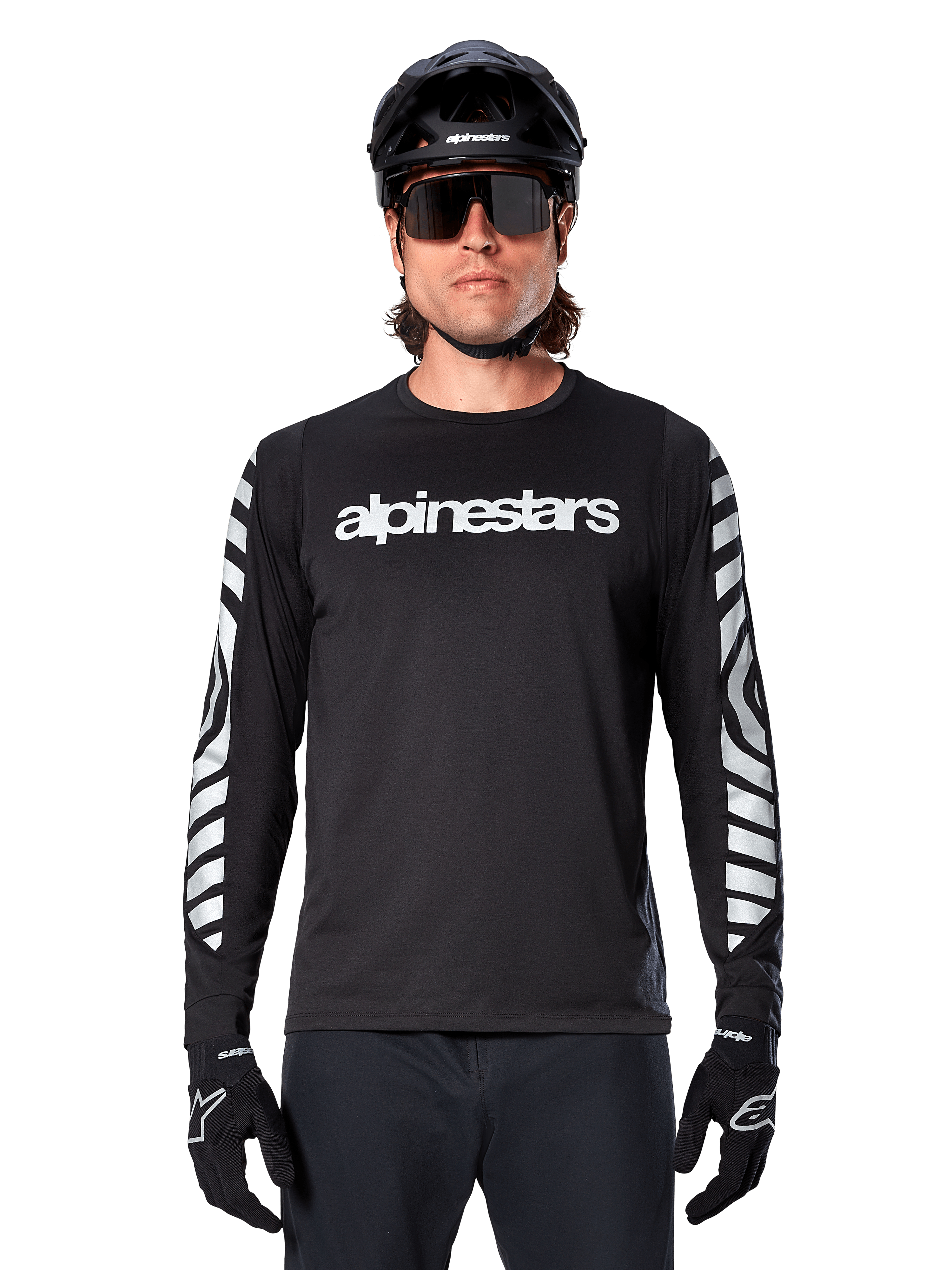 Mężczyzna w koszulce Alpinestars A-Dura Dri Oscar z długim rękawem, kolor Black Reflective, z białym napisem Alpinestars na klatce piersiowej i srebrnymi odblaskowymi grafikami na rękawach, czarny kask MTB, okulary przeciwsłoneczne i czarne rękawiczki, profesjonalna odzież rowerowa