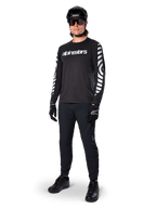 Koszulka Alpinestars A-Dura Dri Oscar z długim rękawem, Black Reflective, z wyraźnym białym napisem Alpinestars na klatce piersiowej i odblaskowymi wzorami graficznymi na rękawach, noszona z czarnymi spodniami MTB, kaskiem i rękawicami, techniczna odzież MTB