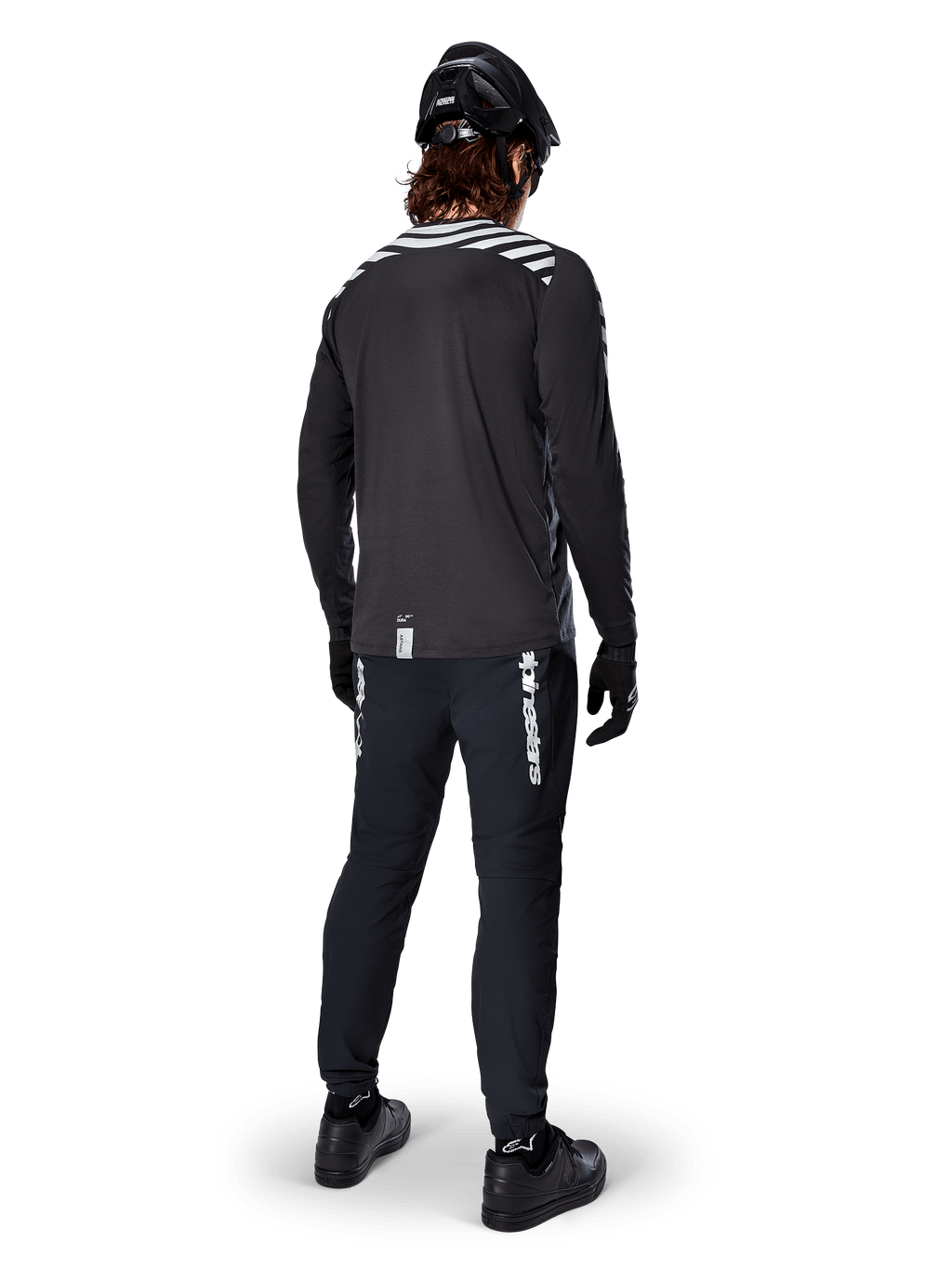 Rowerzysta MTB w koszulce Alpinestars A-Dura Dri Oscar z długim rękawem w kolorze Black Reflective, widok z tyłu, czarne spodnie techniczne, kask i rękawice, odblaskowe paski na ramionach i przedłużony tył do jazdy w terenie.
