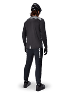 Rowerzysta MTB w koszulce Alpinestars A-Dura Dri Oscar z długim rękawem w kolorze Black Reflective, widok z tyłu, czarne spodnie techniczne, kask i rękawice, odblaskowe paski na ramionach i przedłużony tył do jazdy w terenie.