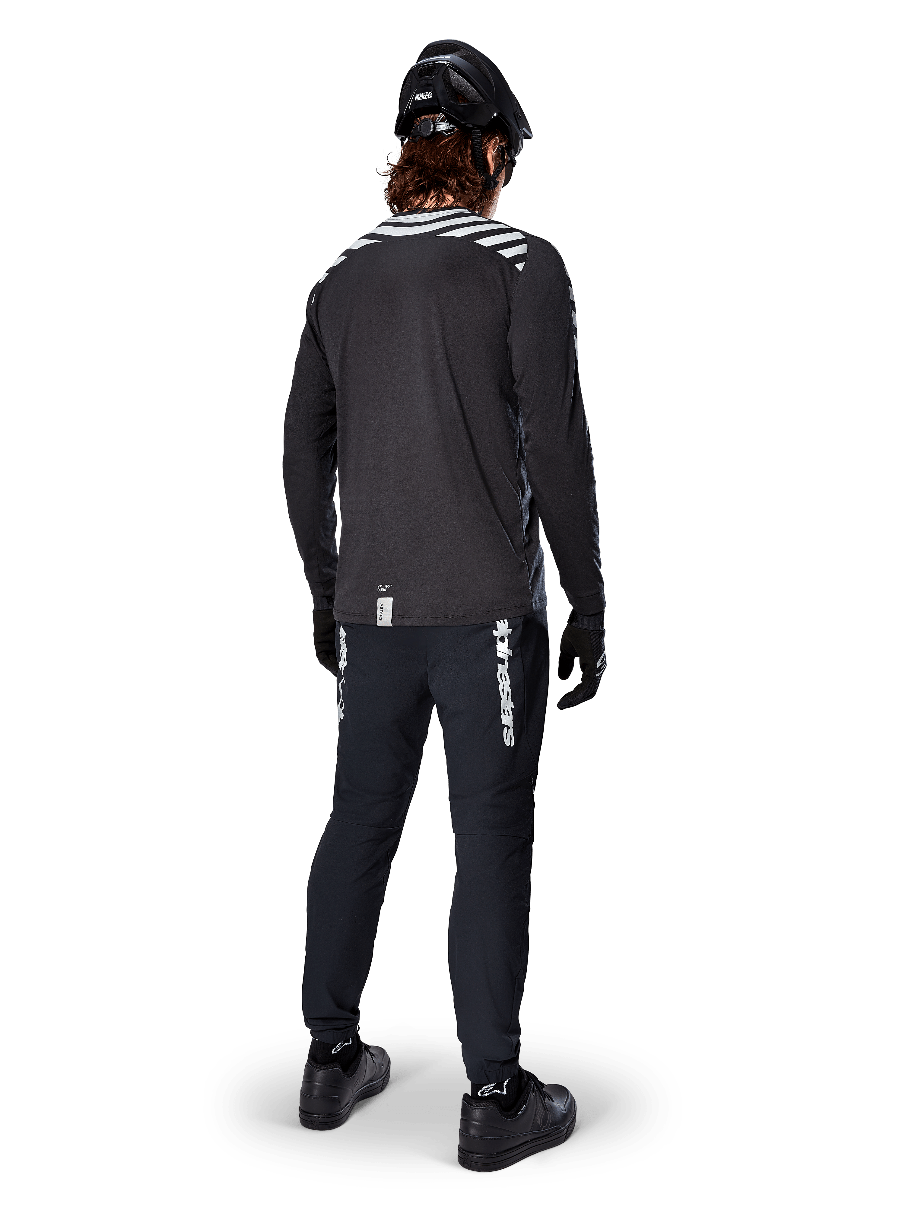 Rowerzysta MTB w koszulce Alpinestars A-Dura Dri Oscar z długim rękawem w kolorze Black Reflective, widok z tyłu, czarne spodnie techniczne, kask i rękawice, odblaskowe paski na ramionach i przedłużony tył do jazdy w terenie.