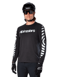 Koszulka z długim rękawem Alpinestars A-Dura Dri Oscar, koszulka rowerowa MTB, czarna odblaskowa, z białym napisem na klatce piersiowej i wyraźnymi odblaskowymi grafikami na rękawach, noszona z czarnym kaskiem i okularami przeciwsłonecznymi, techniczna odzież odprowadzająca wilgoć do jazdy terenowej na rowerze górskim