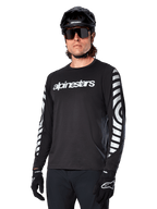 Koszulka z długim rękawem Alpinestars A-Dura Dri Oscar, koszulka rowerowa MTB, czarna odblaskowa, z białym napisem na klatce piersiowej i wyraźnymi odblaskowymi grafikami na rękawach, noszona z czarnym kaskiem i okularami przeciwsłonecznymi, techniczna odzież odprowadzająca wilgoć do jazdy terenowej na rowerze górskim