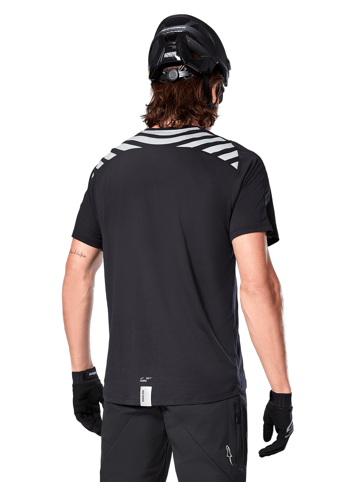 Alpinestars A-Dura Dri Oscar Jersey, koszulka MTB z krótkim rękawem, Black Reflective, widok z tyłu na modelu w kasku rowerowym i rękawiczkach, z odblaskowymi szarymi pasami na ramionach i przedłużonym tyłem, techniczny materiał odprowadzający wilgoć do jazdy na rowerze górskim