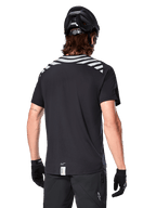 Alpinestars A-Dura Dri Oscar Jersey, koszulka MTB z krótkim rękawem, Black Reflective, widok z tyłu na modelu w kasku rowerowym i rękawiczkach, z odblaskowymi szarymi pasami na ramionach i przedłużonym tyłem, techniczny materiał odprowadzający wilgoć do jazdy na rowerze górskim