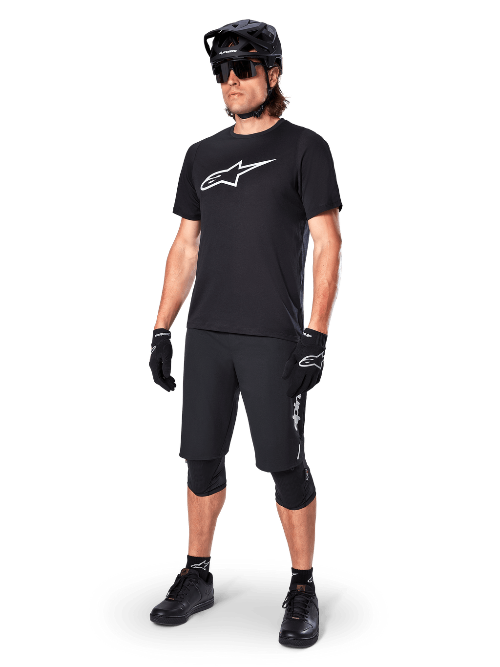 Rowerzysta górski w koszulce Alpinestars A-Dura Dri Oscar Short Sleeve w kolorze Black Reflective, z dużym białym logo na piersi, czarne spodenki MTB, rękawice i kask, techniczna odzież kolarska do jazdy w terenie