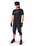 Rowerzysta górski w koszulce Alpinestars A-Dura Dri Oscar Short Sleeve w kolorze Black Reflective, z dużym białym logo na piersi, czarne spodenki MTB, rękawice i kask, techniczna odzież kolarska do jazdy w terenie