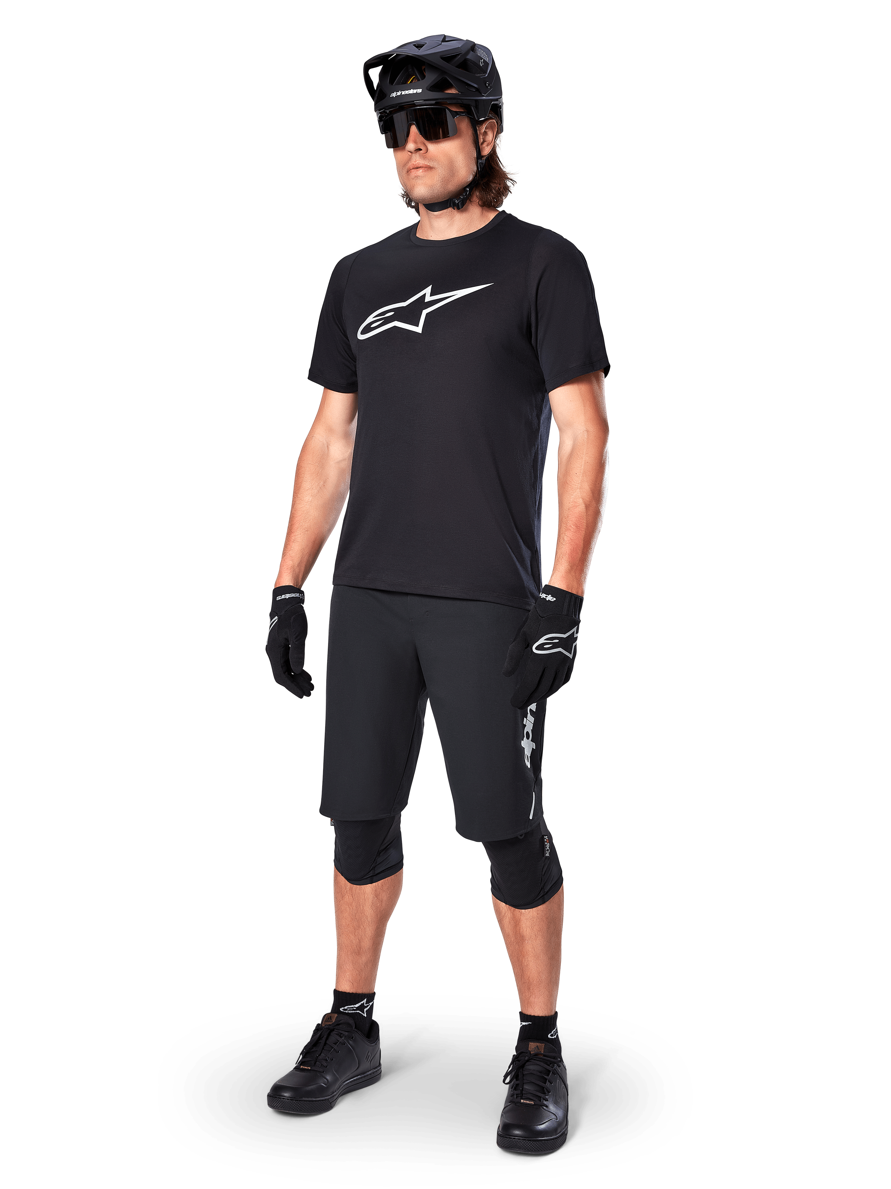 Rowerzysta górski w koszulce Alpinestars A-Dura Dri Oscar Short Sleeve w kolorze Black Reflective, z dużym białym logo na piersi, czarne spodenki MTB, rękawice i kask, techniczna odzież kolarska do jazdy w terenie