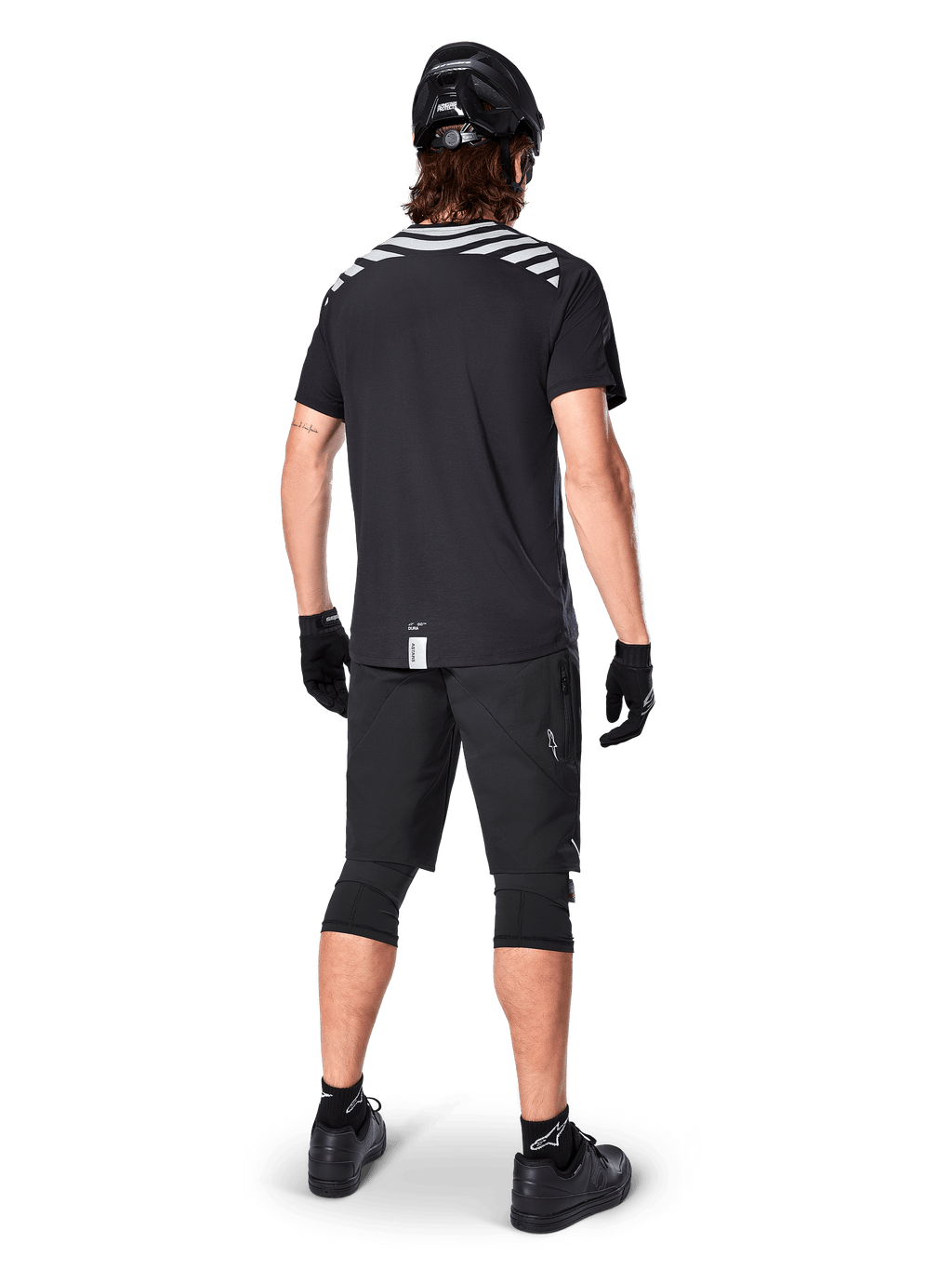 Alpinestars A-Dura Dri Oscar Short Sleeve Jersey, Black Reflective, widok z tyłu rowerzysty ubranego w koszulkę, czarne szorty MTB, kask i ochraniacze na kolana, z odblaskowymi grafikami na ramionach i przedłużonym tyłem do jazdy MTB.