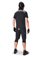 Alpinestars A-Dura Dri Oscar Short Sleeve Jersey, Black Reflective, widok z tyłu rowerzysty ubranego w koszulkę, czarne szorty MTB, kask i ochraniacze na kolana, z odblaskowymi grafikami na ramionach i przedłużonym tyłem do jazdy MTB.