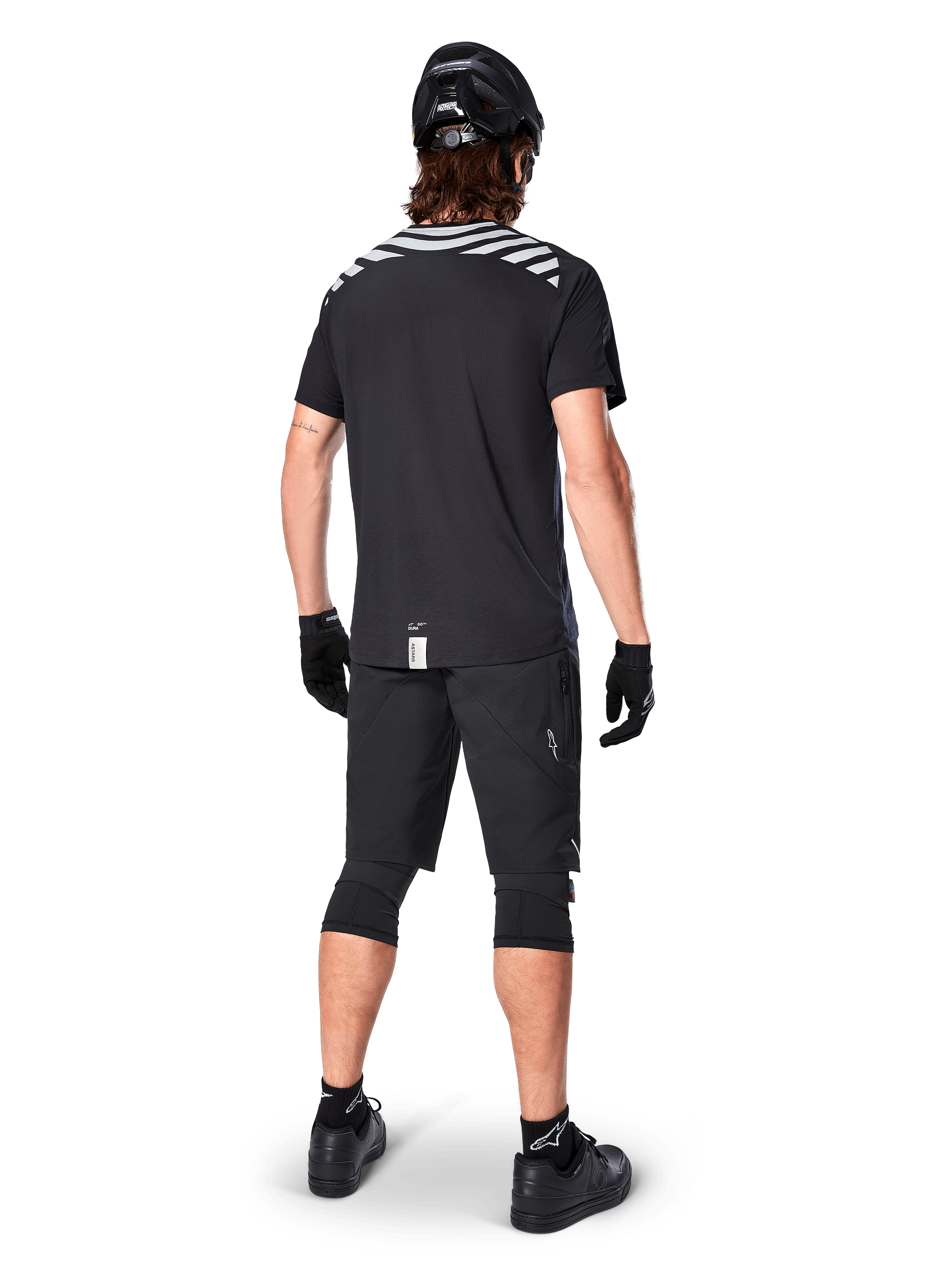Alpinestars A-Dura Dri Oscar Short Sleeve Jersey, Black Reflective, widok z tyłu rowerzysty ubranego w koszulkę, czarne szorty MTB, kask i ochraniacze na kolana, z odblaskowymi grafikami na ramionach i przedłużonym tyłem do jazdy MTB.