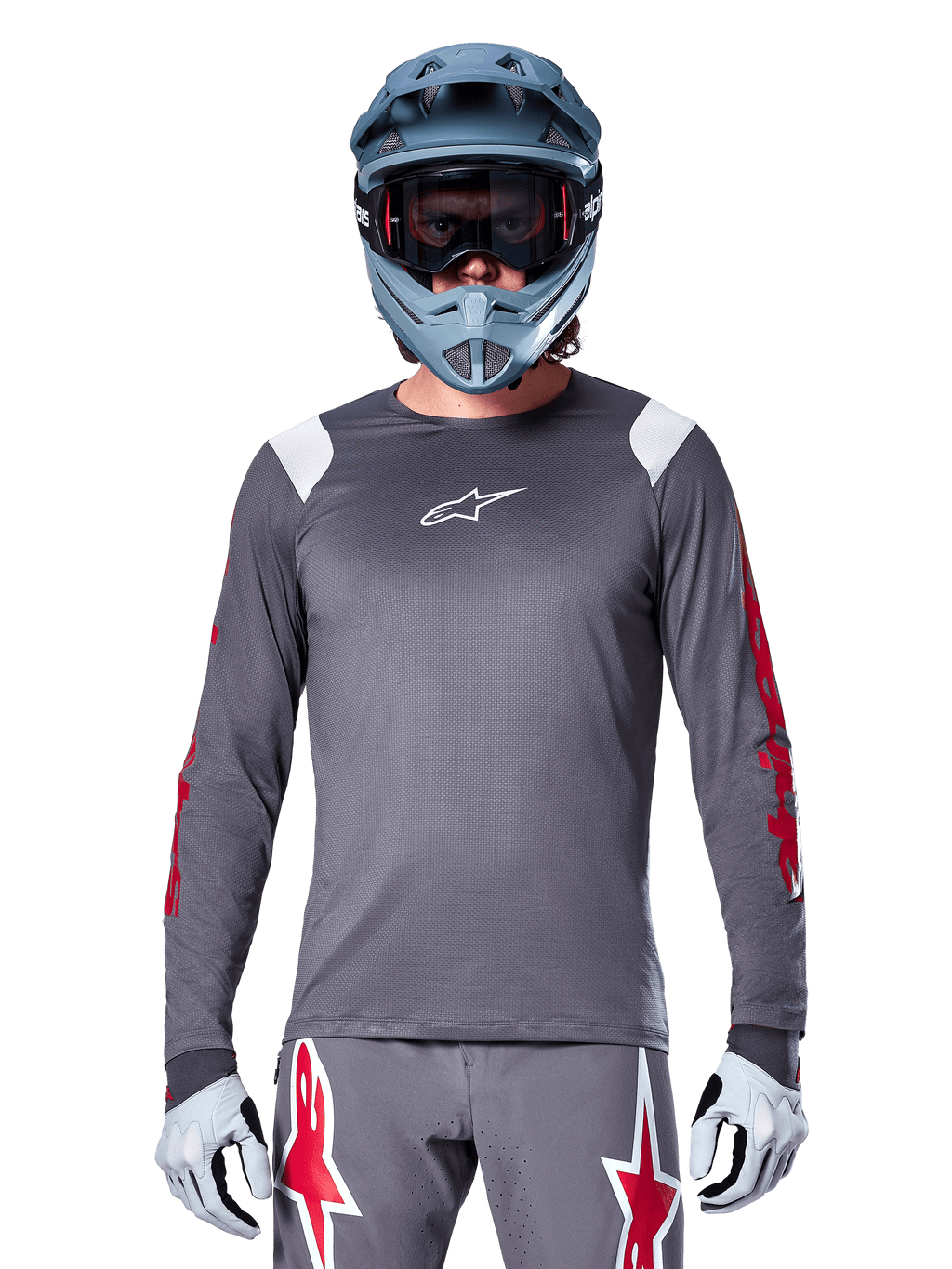Rowerzysta MTB w koszulce Alpinestars A-Supra Astar Long Sleeve, ciemnoszara z czerwonymi grafikami na rękawach i białymi akcentami na ramionach, pasujące szare spodnie z czerwonym logo gwiazdy, niebieski kask full-face i gogle, dopasowany strój do jazdy terenowej