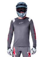 Rowerzysta MTB w koszulce Alpinestars A-Supra Astar Long Sleeve, ciemnoszara z czerwonymi grafikami na rękawach i białymi akcentami na ramionach, pasujące szare spodnie z czerwonym logo gwiazdy, niebieski kask full-face i gogle, dopasowany strój do jazdy terenowej