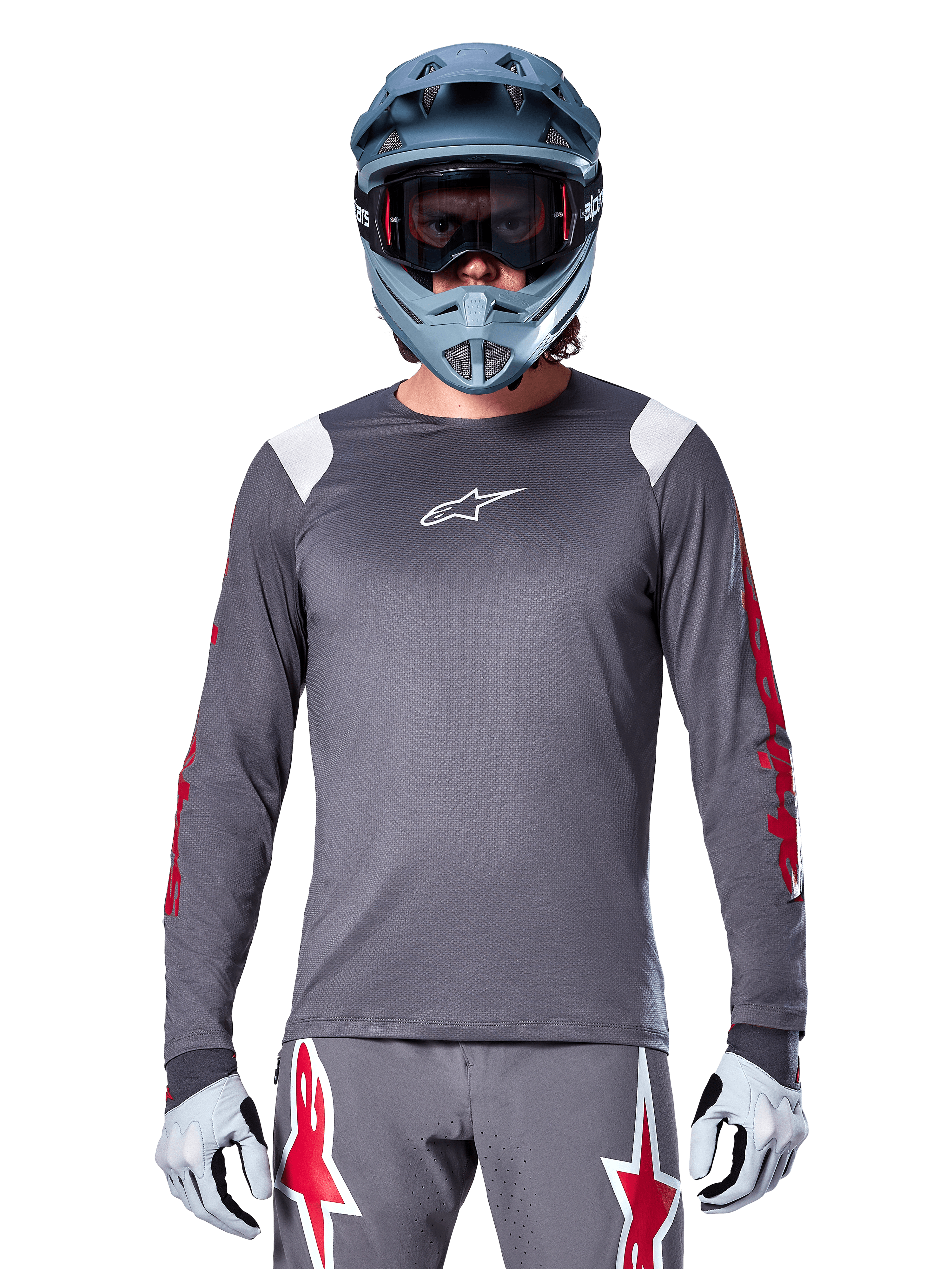 Rowerzysta MTB w koszulce Alpinestars A-Supra Astar Long Sleeve, ciemnoszara z czerwonymi grafikami na rękawach i białymi akcentami na ramionach, pasujące szare spodnie z czerwonym logo gwiazdy, niebieski kask full-face i gogle, dopasowany strój do jazdy terenowej
