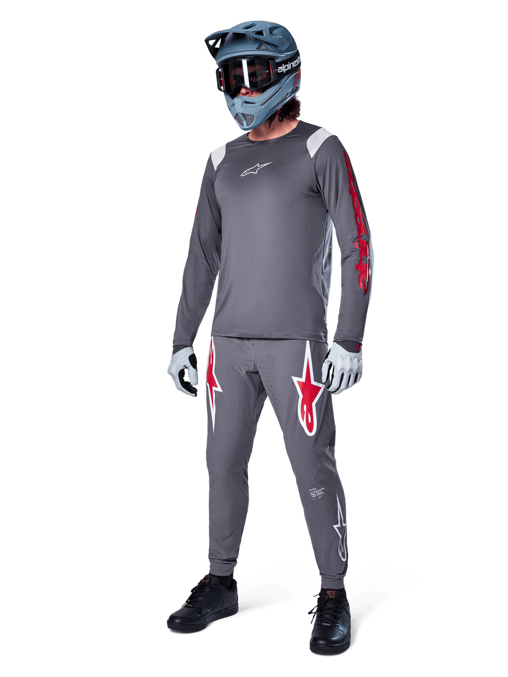 Koszulka z długim rękawem Alpinestars A-Supra Astar w kolorze ciemnoszarym, z czerwonymi logotypami na rękawach i białym logo na piersi, noszona przez rowerzystę w pasujących szarych spodniach, niebiesko-szarym kasku full-face i rękawicach technicznych, zintegrowany zestaw MTB do jazdy grawitacyjnej.