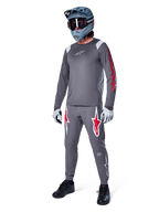 Koszulka z długim rękawem Alpinestars A-Supra Astar w kolorze ciemnoszarym, z czerwonymi logotypami na rękawach i białym logo na piersi, noszona przez rowerzystę w pasujących szarych spodniach, niebiesko-szarym kasku full-face i rękawicach technicznych, zintegrowany zestaw MTB do jazdy grawitacyjnej.