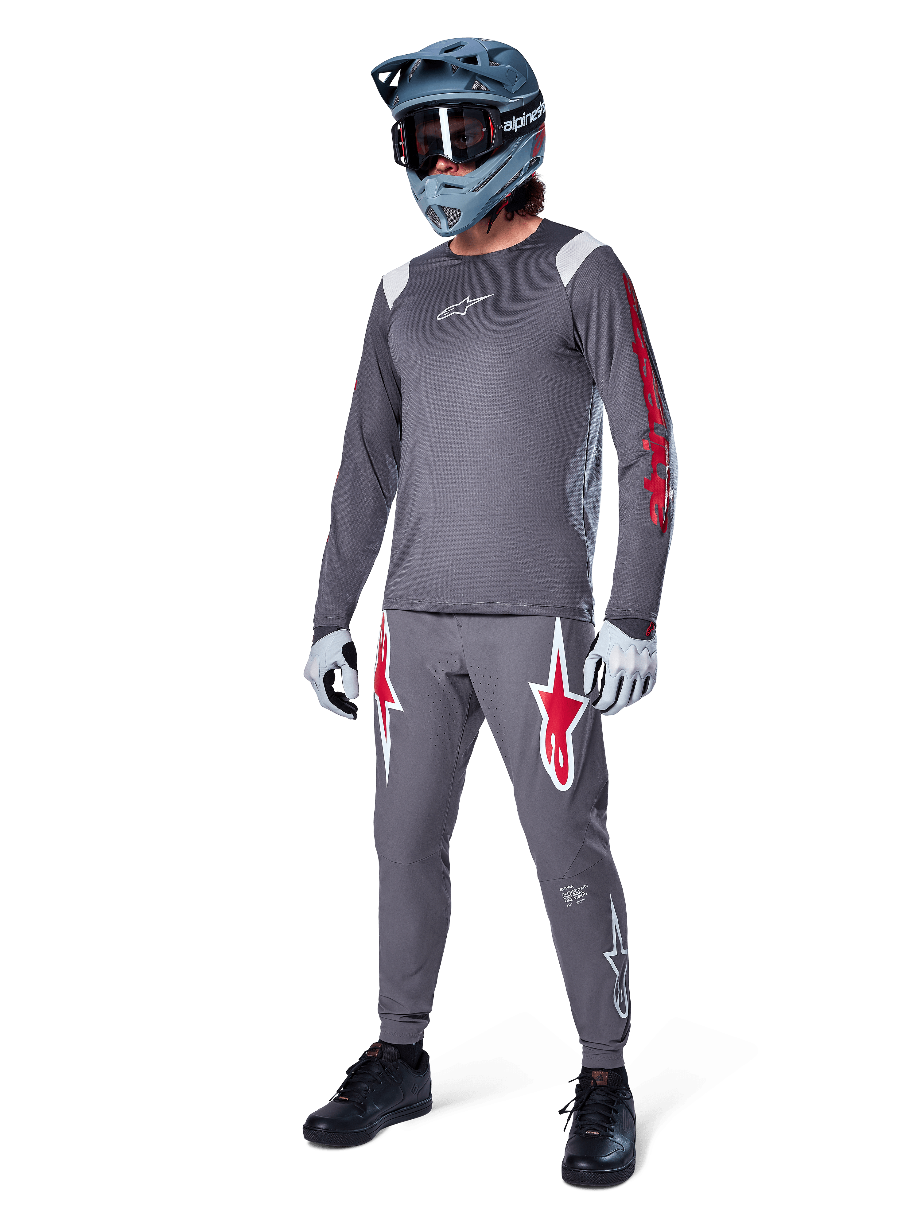 Koszulka z długim rękawem Alpinestars A-Supra Astar w kolorze ciemnoszarym, z czerwonymi logotypami na rękawach i białym logo na piersi, noszona przez rowerzystę w pasujących szarych spodniach, niebiesko-szarym kasku full-face i rękawicach technicznych, zintegrowany zestaw MTB do jazdy grawitacyjnej.