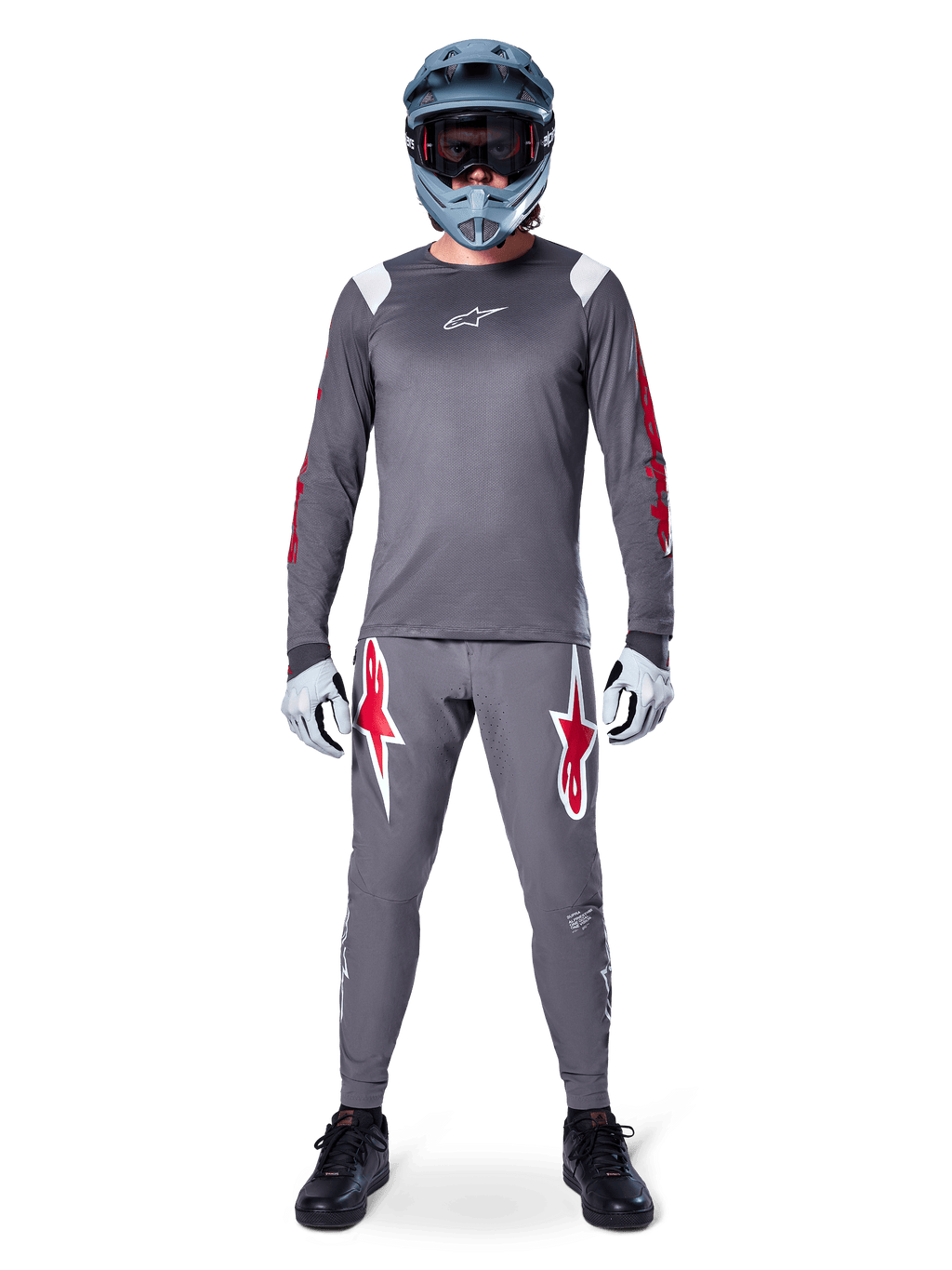 Rowerzysta MTB w koszulce Alpinestars A-Supra Astar Long Sleeve, ciemnoszara z czerwonymi akcentami i białym logo na piersi, pasujące szare spodnie z dużymi grafikami czerwonej gwiazdy, niebieski kask full-face z goglami, jasnoszare rękawiczki i czarne buty, profesjonalny zestaw odzieży na rower górski