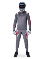 Rowerzysta MTB w koszulce Alpinestars A-Supra Astar Long Sleeve, ciemnoszara z czerwonymi akcentami i białym logo na piersi, pasujące szare spodnie z dużymi grafikami czerwonej gwiazdy, niebieski kask full-face z goglami, jasnoszare rękawiczki i czarne buty, profesjonalny zestaw odzieży na rower górski