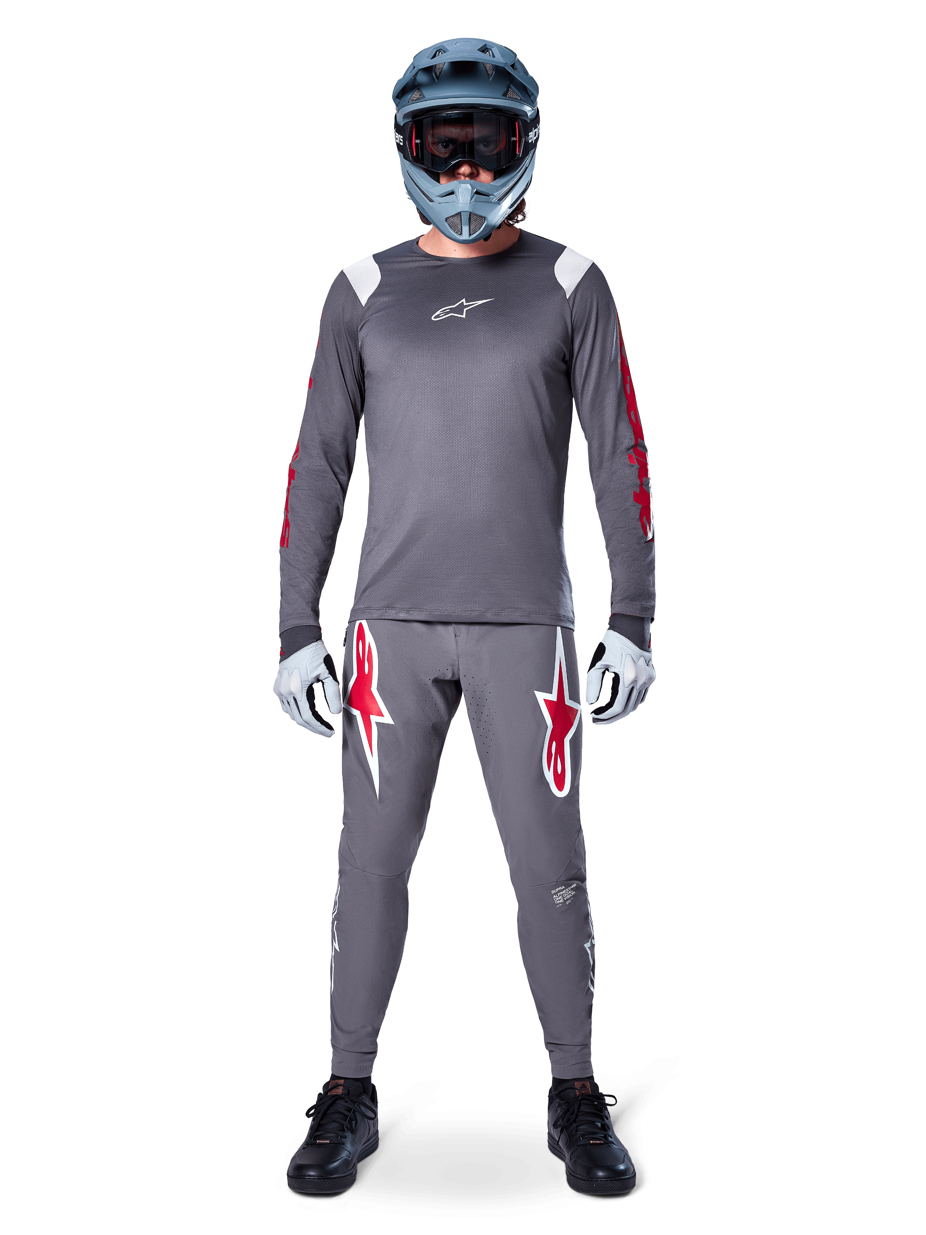 Rowerzysta MTB w koszulce Alpinestars A-Supra Astar Long Sleeve, ciemnoszara z czerwonymi akcentami i białym logo na piersi, pasujące szare spodnie z dużymi grafikami czerwonej gwiazdy, niebieski kask full-face z goglami, jasnoszare rękawiczki i czarne buty, profesjonalny zestaw odzieży na rower górski
