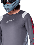 Alpinestars A-Supra Astar Long Sleeve Jersey, koszulka z długim rękawem, ciemnoszara z białymi panelami na ramionach i czerwonymi grafikami na rękawach, noszona z jasnym niebiesko-szarym kaskiem full-face, profesjonalna odzież do kolarstwa górskiego
