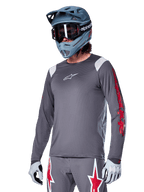 Alpinestars A-Supra Astar Jersey z długim rękawem, koszulka rowerowa MTB, ciemnoszara z czerwonymi i białymi akcentami, sportowy krój, noszona z pasującymi szarymi spodniami z grafiką czerwonych i białych gwiazd, kompletny strój do kolarstwa terenowego z kaskiem full-face i rękawicami