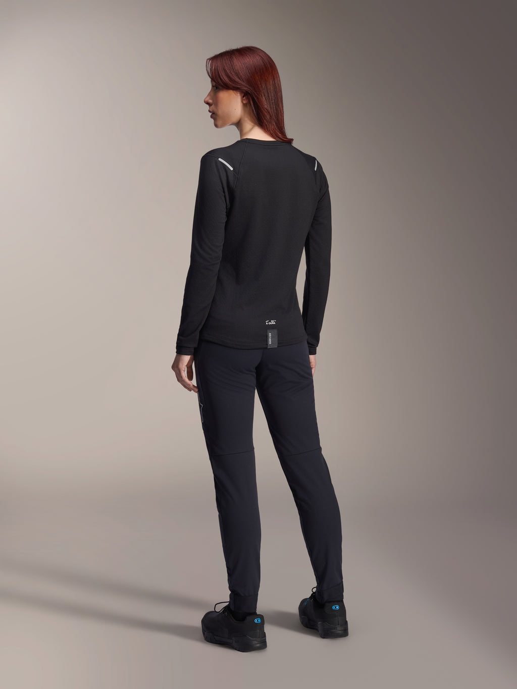 Alpinestars Stella A-Dura Thermal Jersey, damska koszulka MTB z długim rękawem w kolorze czarnym, widok z tyłu pokazujący odblaskowe akcenty na ramionach i przedłużony tył, noszona z czarnymi spodniami i butami, profesjonalna odzież do kolarstwa górskiego.