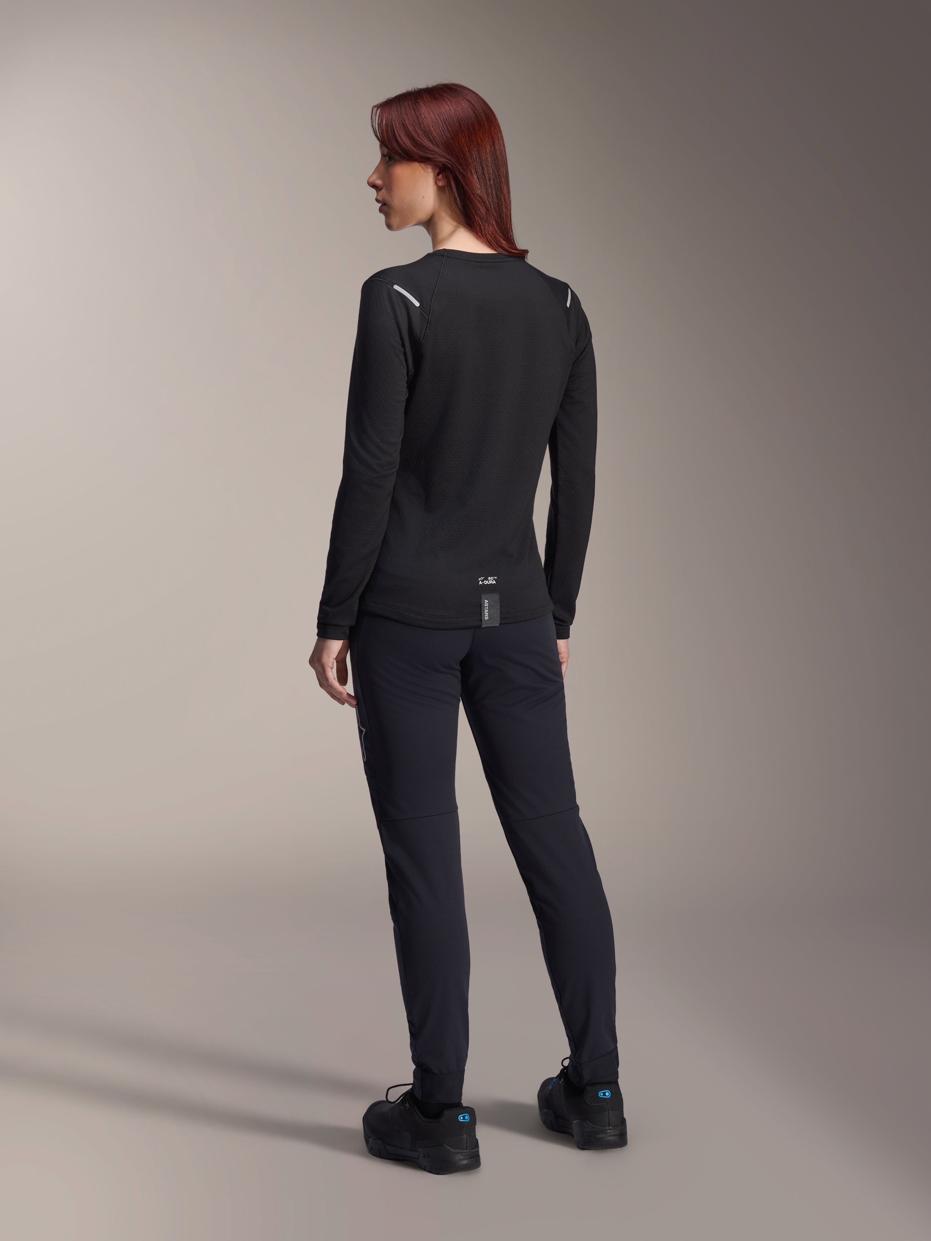 Alpinestars Stella A-Dura Thermal Jersey, damska koszulka MTB z długim rękawem w kolorze czarnym, widok z tyłu pokazujący odblaskowe akcenty na ramionach i przedłużony tył, noszona z czarnymi spodniami i butami, profesjonalna odzież do kolarstwa górskiego.