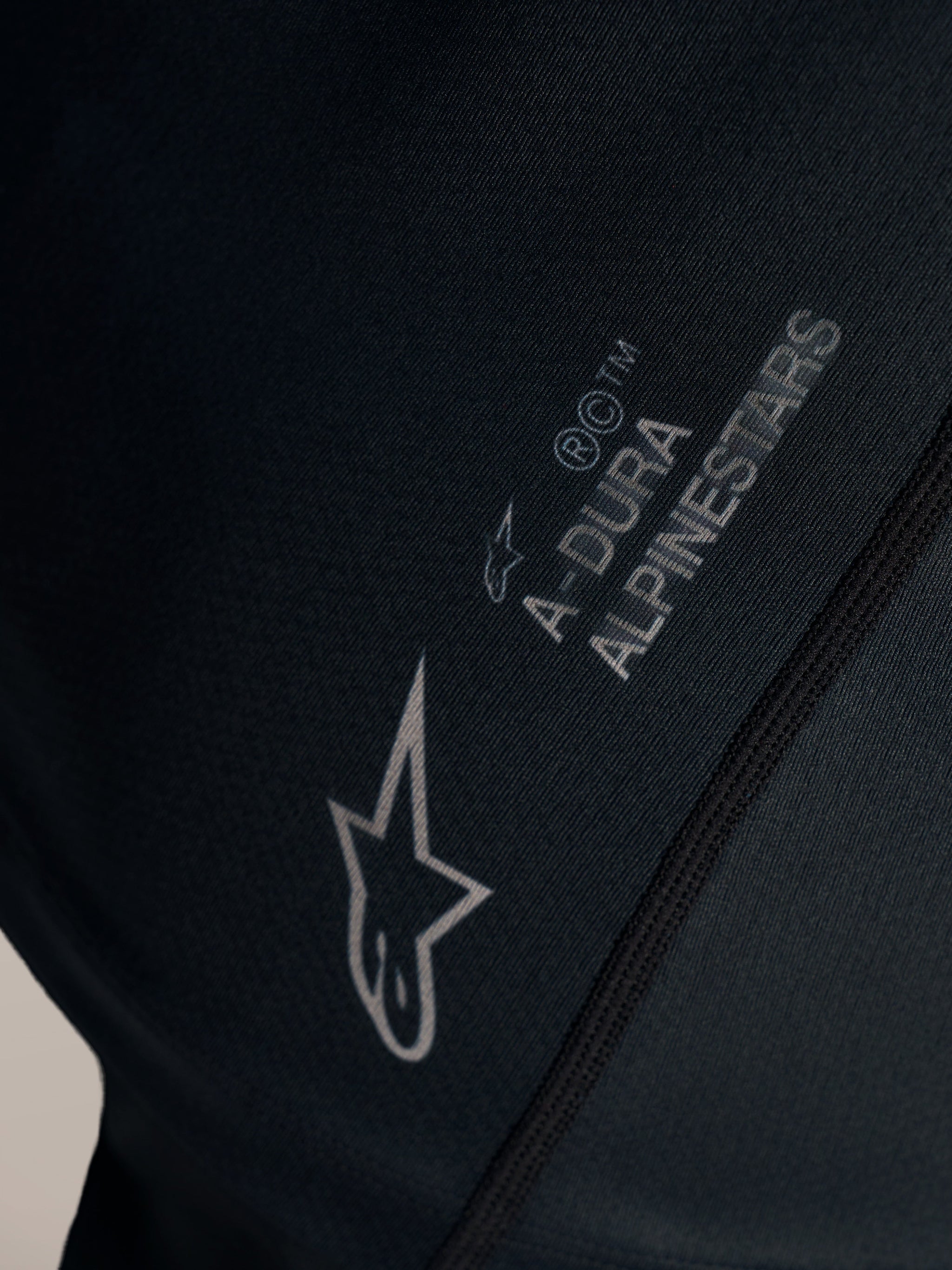 Czarna koszulka Stella A-Dura Ride, zbliżenie na białe logo Alpinestars i branding A-DURA na tkaninie żakardowej.