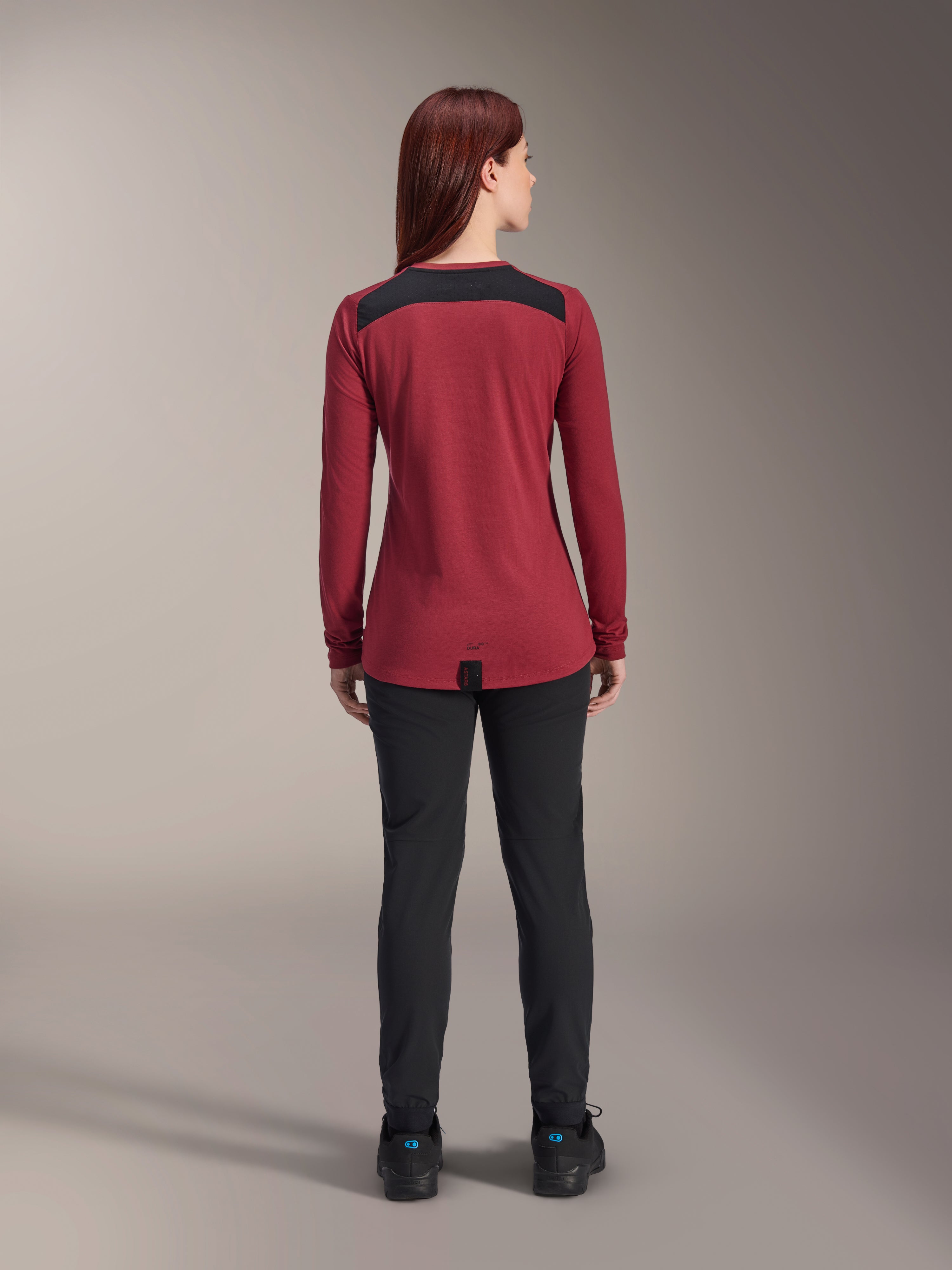 Alpinestars Stella A-Dura Dri Astar Long Sleeve Jersey, damska koszulka z długim rękawem, bordowa z czarnymi siatkowymi panelami na ramionach, techniczna odzież MTB widoczna od tyłu z czarnymi spodniami, wydłużony tył zapewniający osłonę podczas jazdy