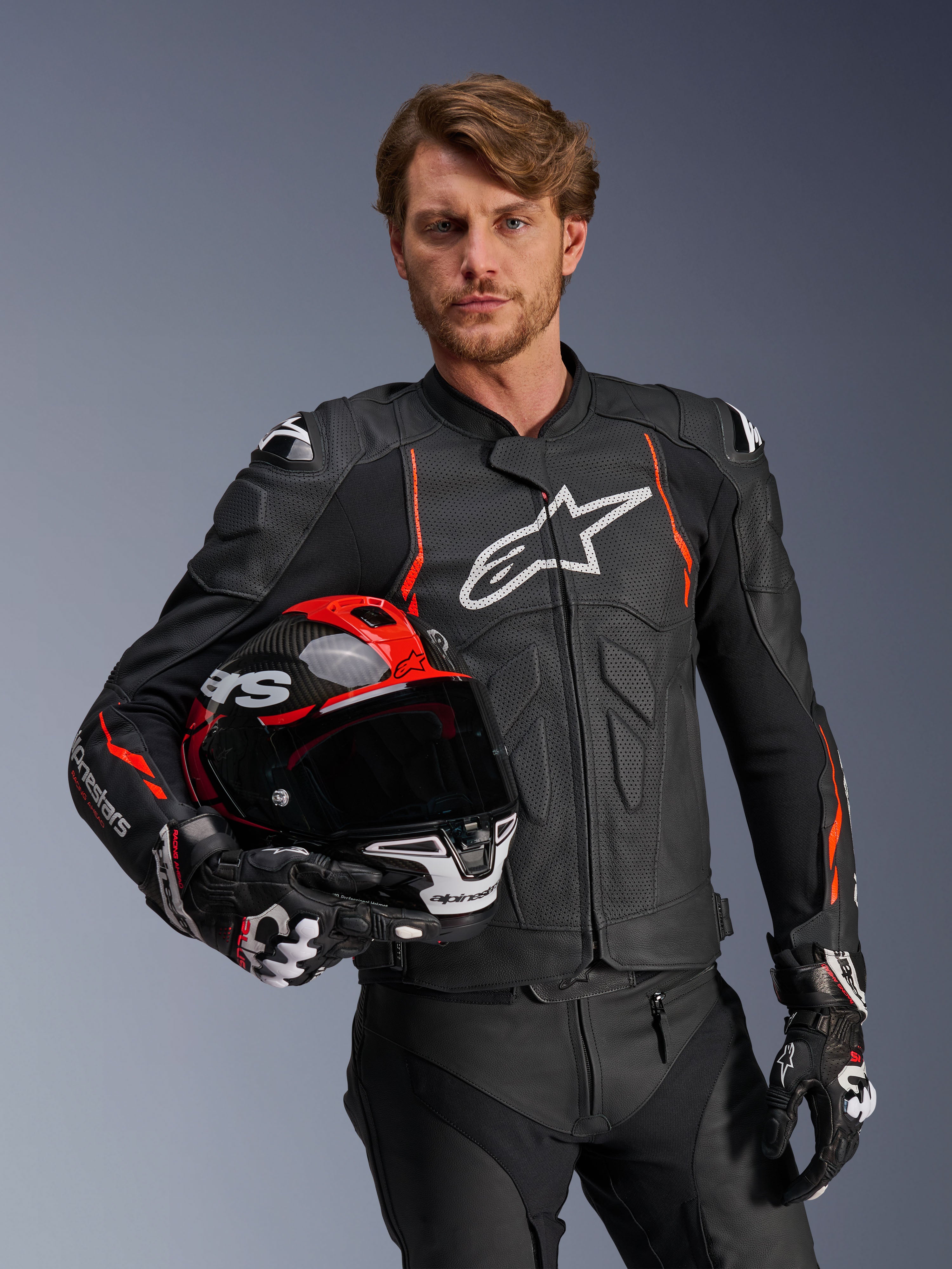 Skórzana kurtka Alpinestars Dusk Airflow, czarna z akcentami Red Fluo, noszona przez modela trzymającego czerwono-czarny kask motocyklowy, z perforowaną skórą dla maksymalnej wentylacji i białym logo Alpinestars, sportowa odzież motocyklowa