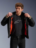 Mężczyzna w czarnej kurtce Alpinestars Ignite Air z siatkowym korpusem i jaskrawopomarańczową odpinaną wiatroodporną podszewką, czarny t-shirt z logo Astars, czarne spodnie, współczesny miejski styl motocyklowy